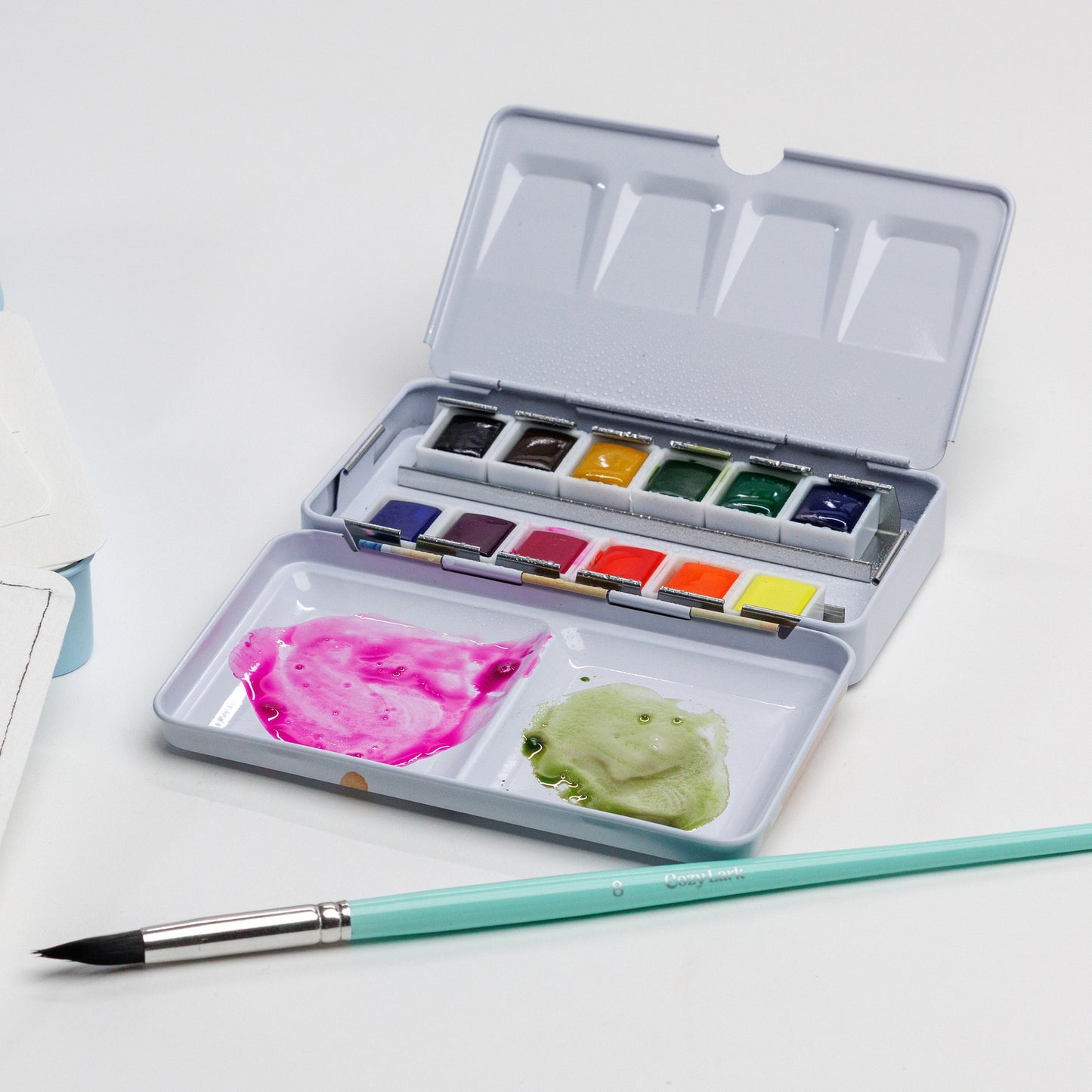 Cozy Lark 12-color watercolor palette with bright paints and a brush on white background