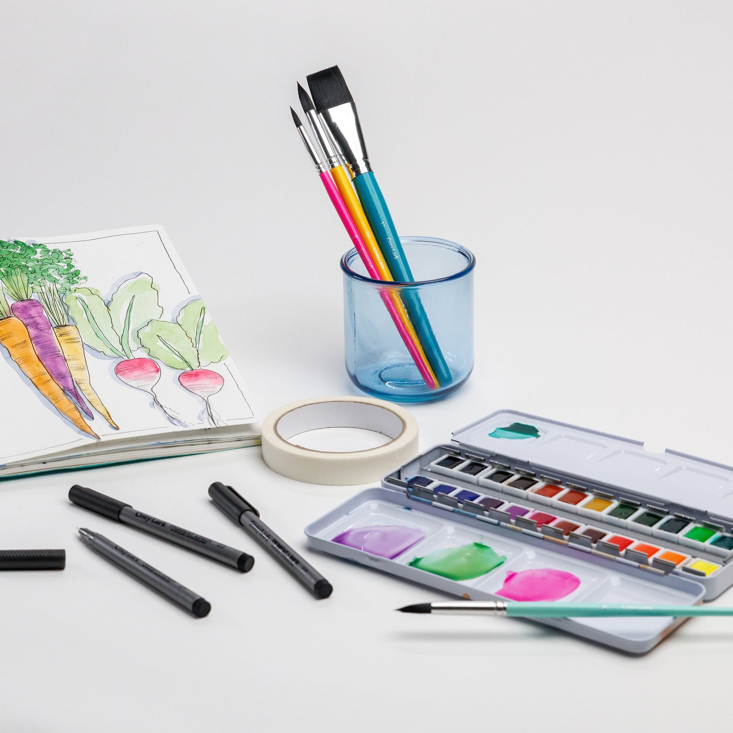 Cozy Lark watercolor sketchbook with brushes, pens, artist tape, and paint palette laid out with a vegetable illustration in the practice book
