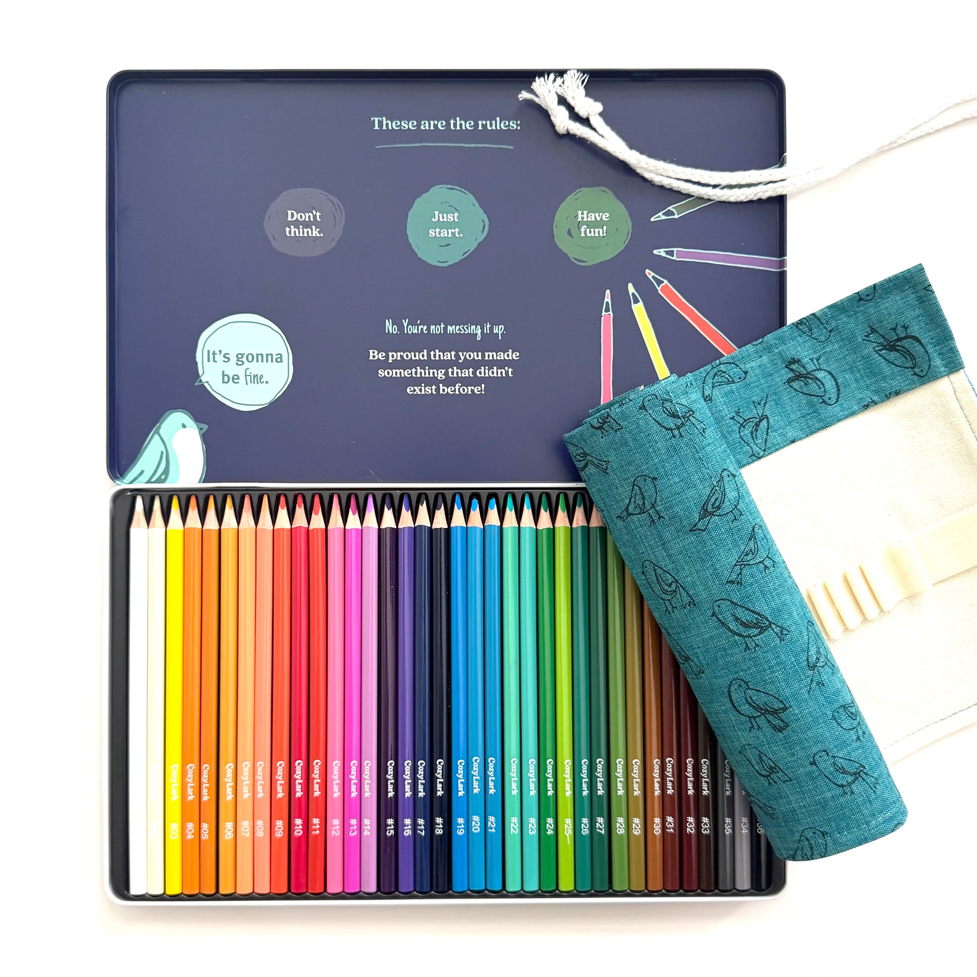 A 24-color pencil set in a dark blue metal tin, shown open with the lid displaying rules and illustrations. A teal fabric pencil pouch with a bird pattern rests on the right side of the tin.