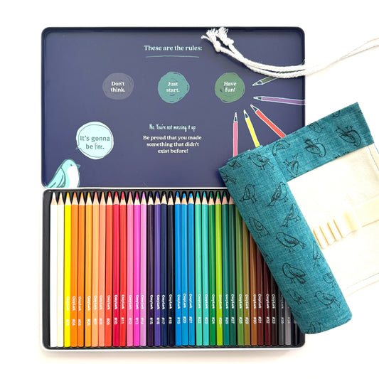 A 24-color pencil set in a dark blue metal tin, shown open with the lid displaying rules and illustrations. A teal fabric pencil pouch with a bird pattern rests on the right side of the tin.
