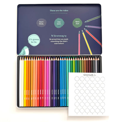 A metal pencil set in an open tin with a navy printed lid, shown alongside a sheet of blank circle stickers. The pencils are arranged in a rainbow of colors, and the lid features playful text and illustrations.