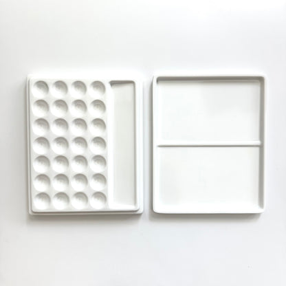 A white ceramic paint palette shown open, displaying a left side with multiple circular wells and a right side with two larger square pans divided by a horizontal line, set against a plain white background.