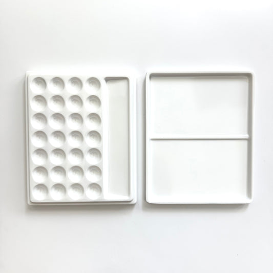 A white ceramic paint palette shown open, displaying a left side with multiple circular wells and a right side with two larger square pans divided by a horizontal line, set against a plain white background.