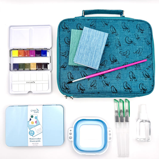 Art supply set with watercolor paints, brushes, postcards, accordion books and travel case on a white background