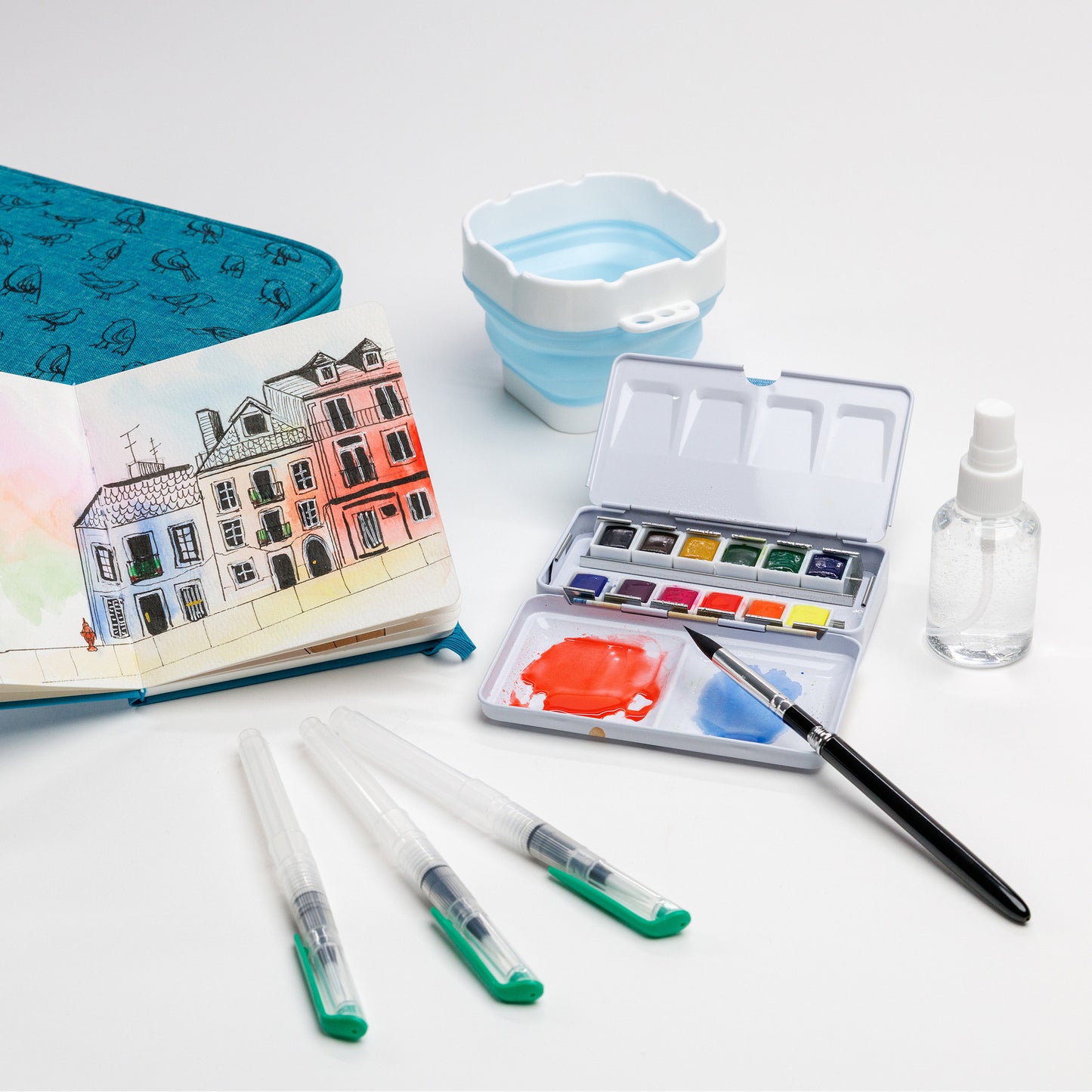 Cozy Lark travel watercolor kit with painted cityscape in sketchbook, collapsible water cup, open palette, three water brushes, and spray bottle on white background