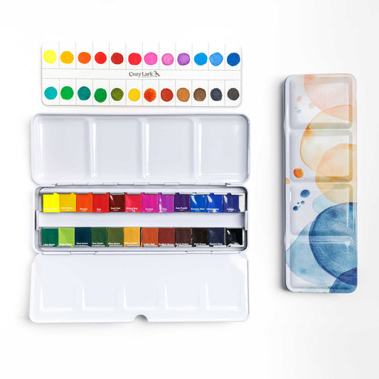 24-color watercolor paint set with stickers on paints, painted swatch cad, and closed tin cover with watercolor circles on a white background