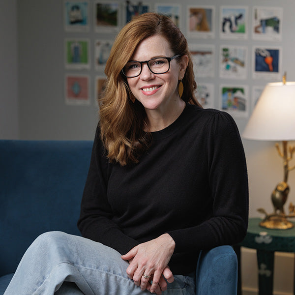Andrea Nelson sitting on a blue sofa with watercolor paintings on the wall behind her. 