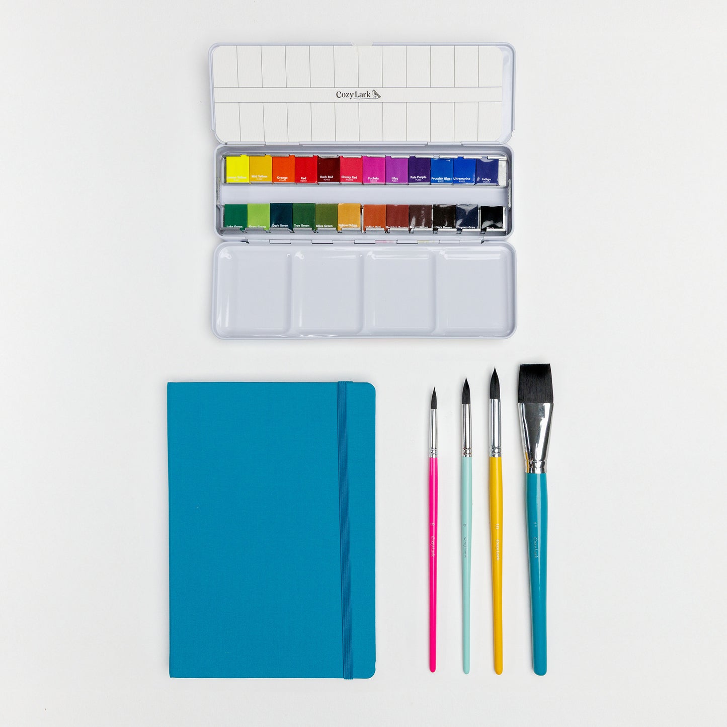 Flat lay of Cozy Lark watercolor palette, artist tape, pigment liners, brushes, and dark teal sketchbook on white background