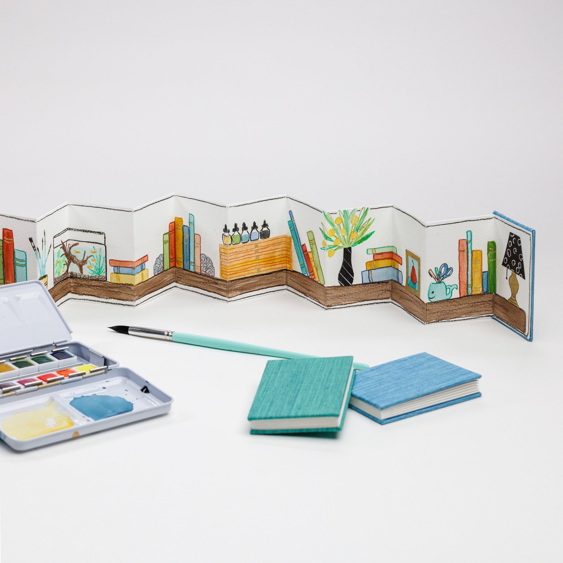 Example of painted mini accordion book with stacked set of mini accordion books, 12-color water color palette, and Size 8 round brush in the foreground