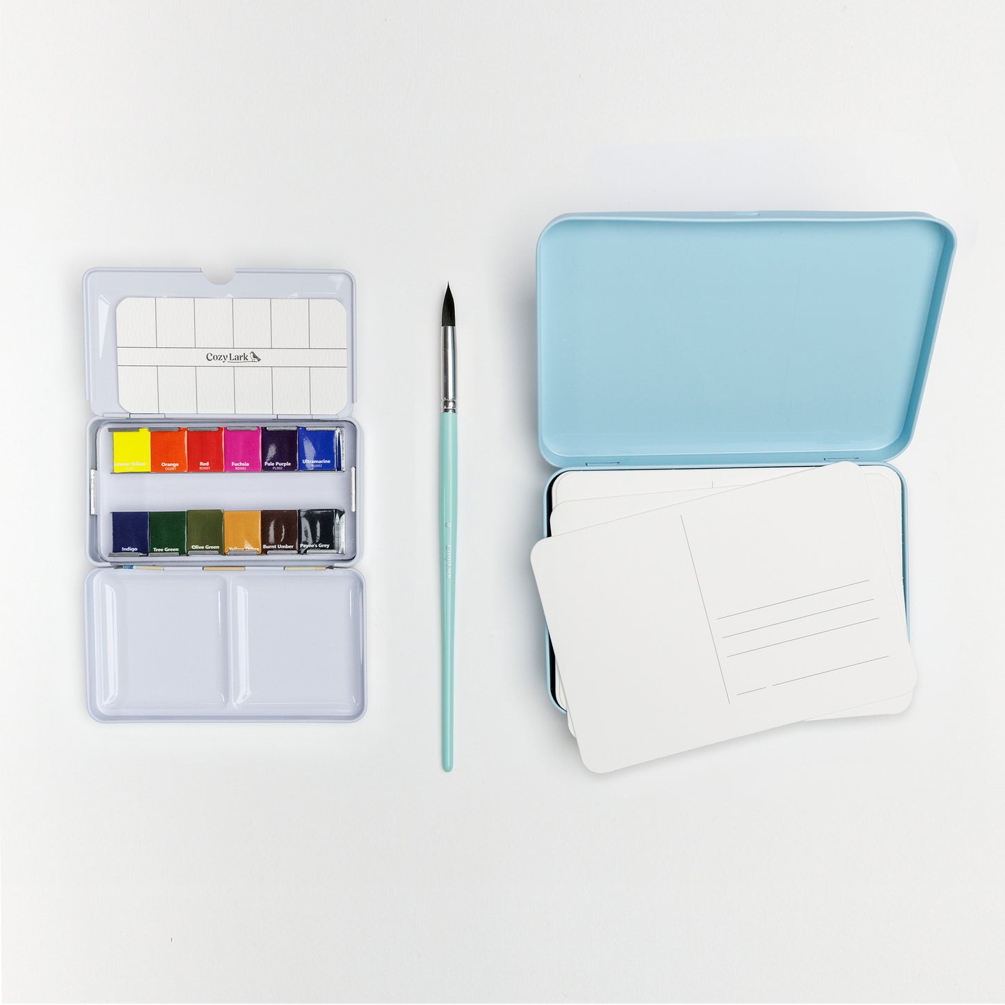 Flat lay of Cozy Lark postcard painting bundle with mini watercolor palette, mint brush, blue tin, and stack of blank watercolor postcards on white background
