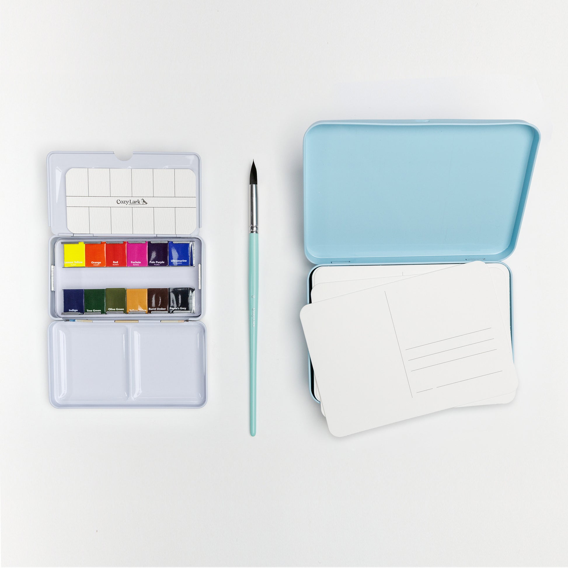 Flat lay of Cozy Lark postcard painting bundle with mini watercolor palette, mint brush, blue tin, and stack of blank watercolor postcards on white background