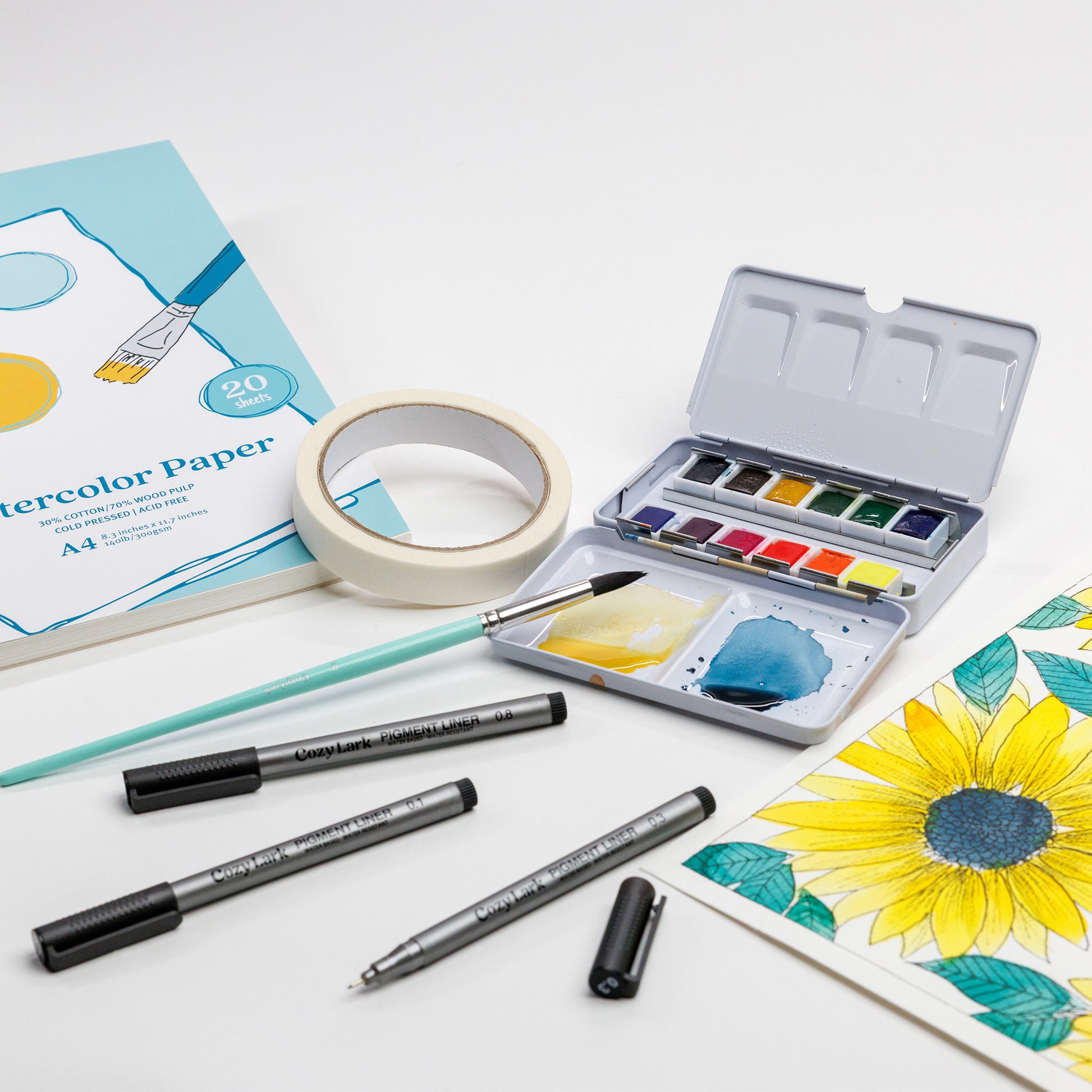 Cozy Lark watercolor starter setup with paper pad, artist  tape, open 12-color palette, Size 8 Round brush, pigment liners, and sunflower painting in progress