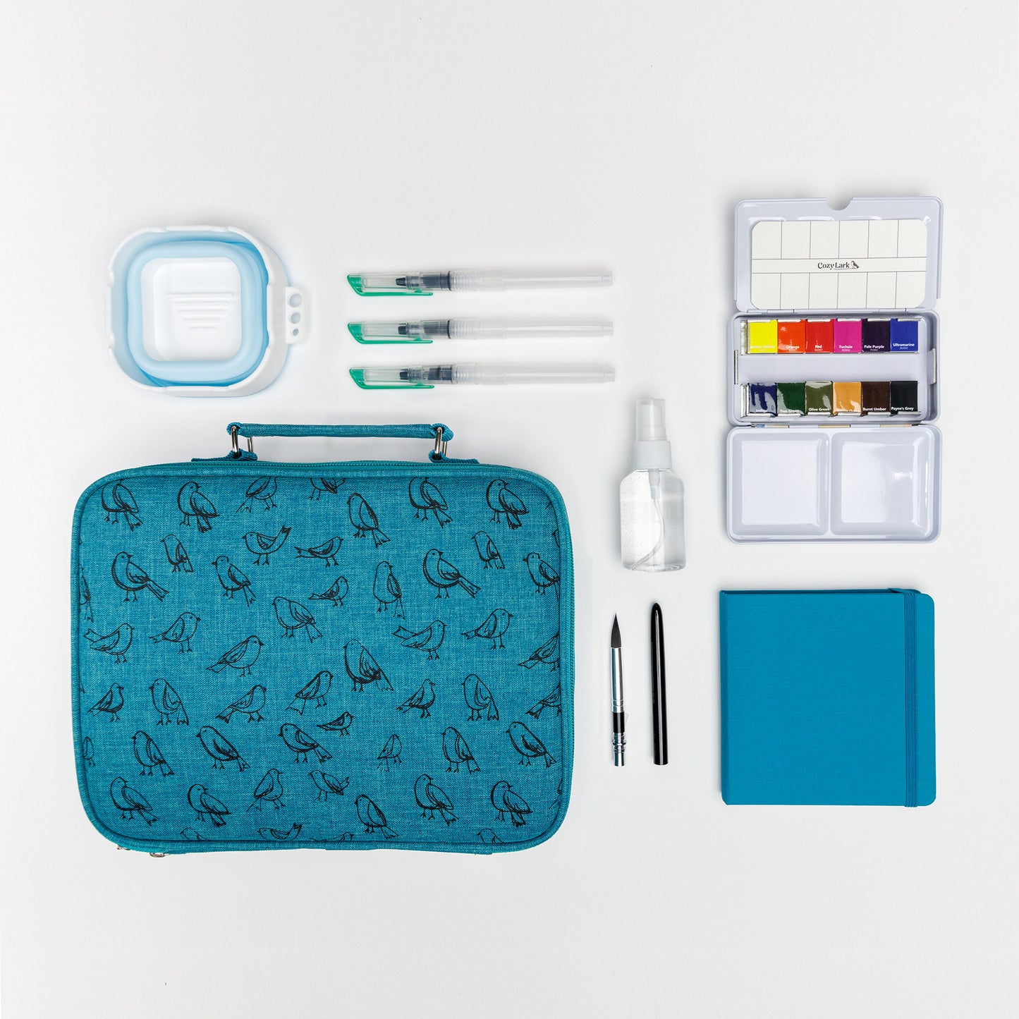 Flat lay of Cozy Lark travel watercolor bundle with teal bird case, mini palette, collapsible water cup, three water brushes, refillable spray bottle, compact brush, and teal travel sketchbook on white background