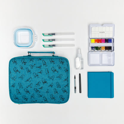 Flat lay of Cozy Lark travel watercolor bundle with teal bird case, mini palette, collapsible water cup, three water brushes, refillable spray bottle, compact brush, and teal travel sketchbook on white background
