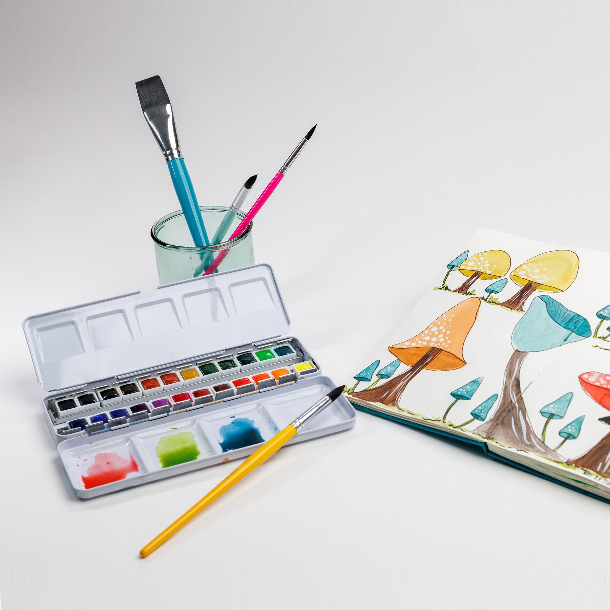Open watercolor palette, three brushes in a glass, and Cozy Lark sketchbook spread with colorful mushroom illustration on white background