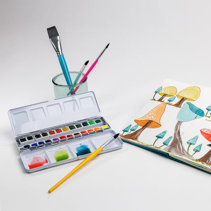 Open watercolor palette, three brushes in a glass, and Cozy Lark sketchbook spread with colorful mushroom illustration on white background